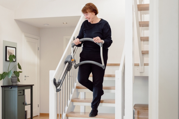 Ältere Frau steigt mit einer mobilen Treppensteighilfe eine Holztreppe im Wohnhaus hinunter.
